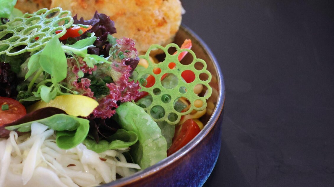 Ein bunter Salat mit verschiedenen Blattsalaten, Gemüse und dekorativen essbaren Pflanzen. Im Hintergrund sind knusprige Mini-Frikadellen zu sehen. | © Gerhard Wolkersdorfer