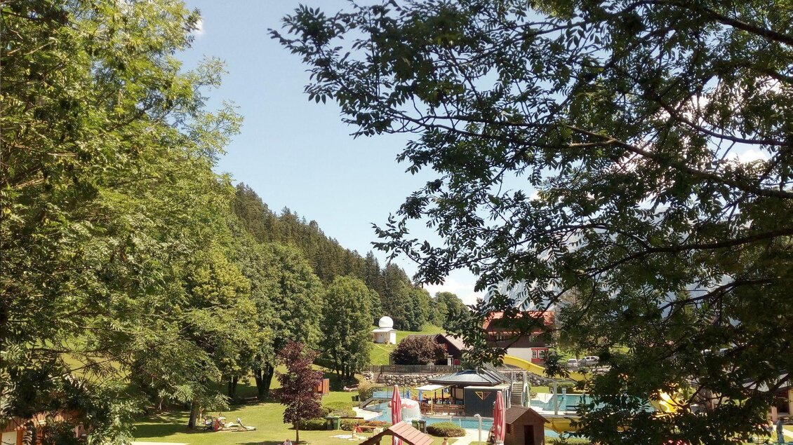 Eine grüne Parklandschaft mit Bäumen und Spielbereichen. Im Hintergrund sind Schwimmbecken und Spielplätze sichtbar. | © Johanna Provatopoulos