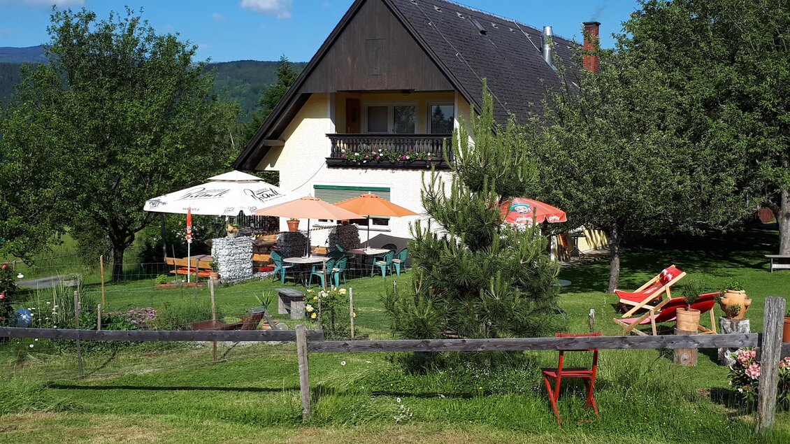 Ein gemütliches Haus mit einem großen Garten und mehreren Sitzbereichen. Umgeben von Bäumen und Sonnenschirmen lädt die Szenerie zum Entspannen ein. | © Gregorhansl