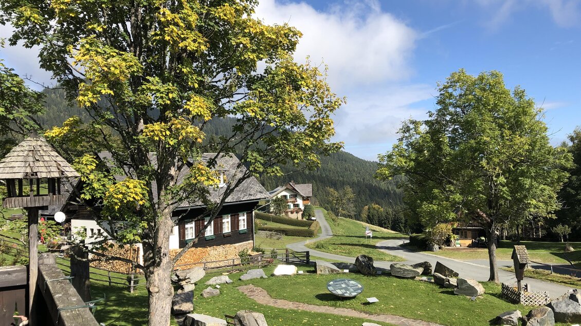 Eine ruhige Landschaft mit einem grünen Garten, Bäumen und kleinen Steinen. Im Hintergrund sind traditionelle Häuser und sanfte Hügel zu sehen. | © Alpenasthof Glashütten