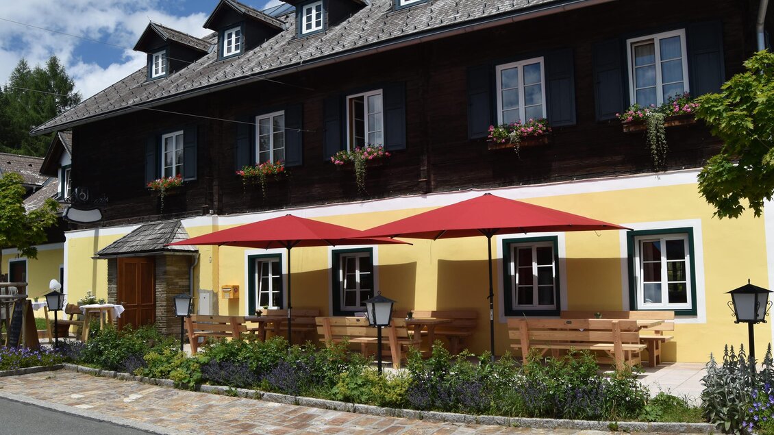 Ein charmantes Gebäude mit gelber Fassade und schwarzen Holzelementen. Draußen stehen Sonnenschirme und Sitzgelegenheiten in einer angenehmen Umgebung. | © Alpenasthof Glashütten