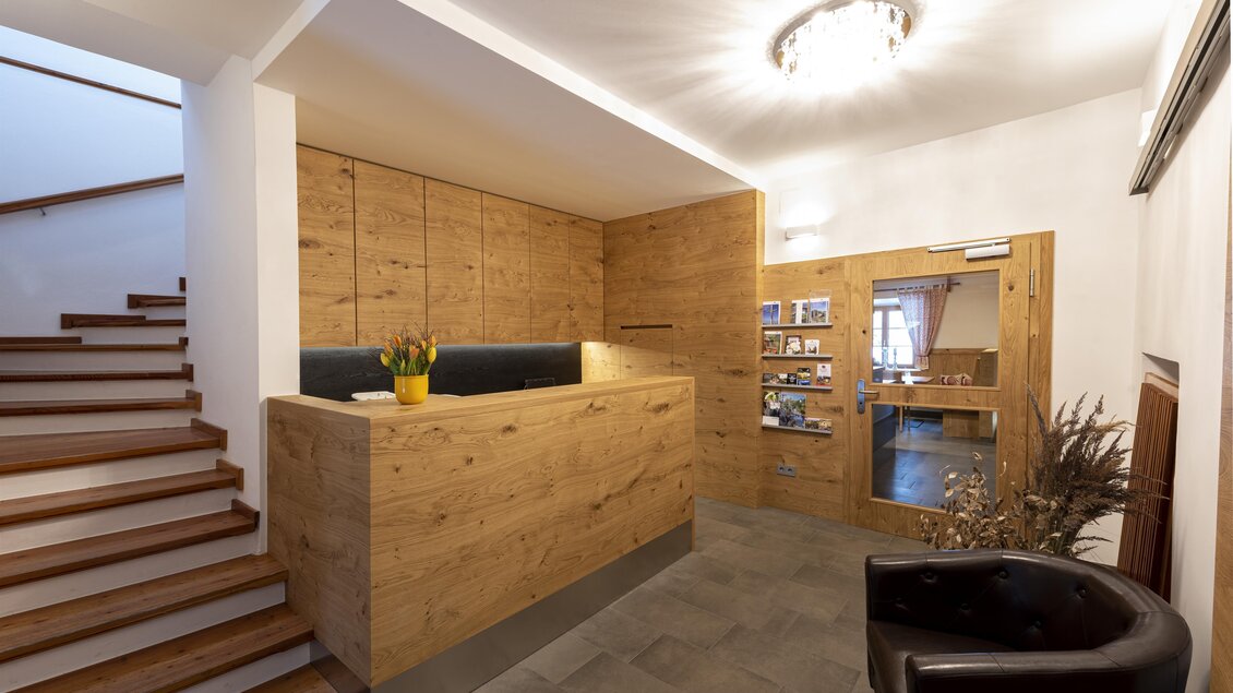 Ein eleganter Empfangsbereich mit Holzverkleidung und einer modernen Theke. Links führt eine Treppe nach oben, und rechts steht ein gemütlicher Sessel. | © Mareike Claar