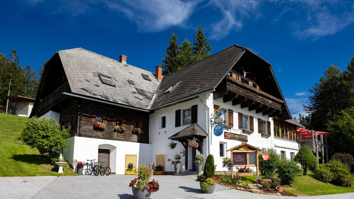 Ein malerisches Gasthaus im alpinen Stil umgeben von grüner Landschaft. Der Himmel ist klar und blau, was eine einladende Atmosphäre schafft. | © TV Region Graz-Harry Schiffer