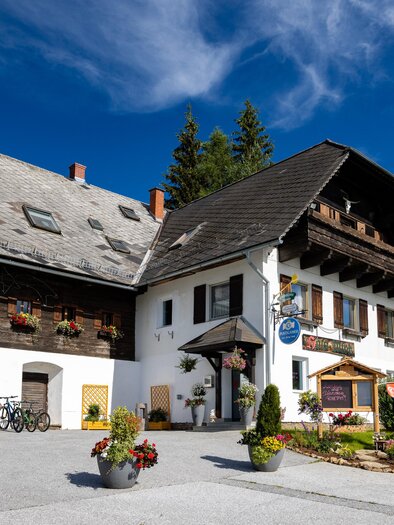 Ein malerisches Gasthaus im alpinen Stil umgeben von grüner Landschaft. Der Himmel ist klar und blau, was eine einladende Atmosphäre schafft. | © TV Region Graz-Harry Schiffer