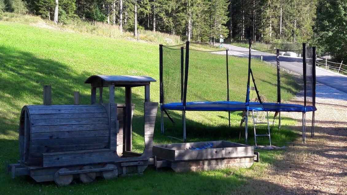 Eine Holzeisenbahn und ein Trampolin stehen auf einer grünen Wiese. Im Hintergrund sind Bäume und ein Weg zu sehen. | © Alpengasthof Hoiswirt