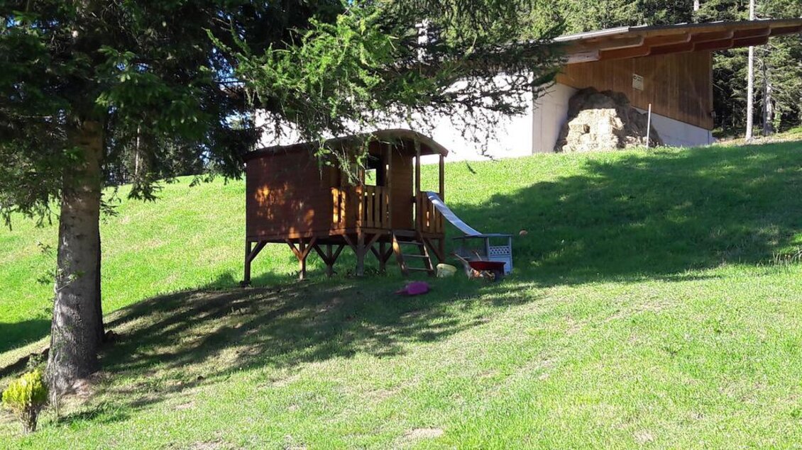 Ein kleines Holzhaus auf einer grünen Wiese mit einer Rutsche. Im Hintergrund stehen Bäume und ein weiteres Gebäude. | © Alpengasthof Hoiswirt