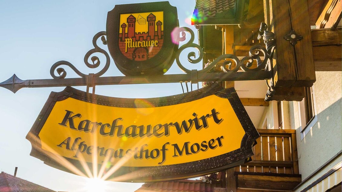 Ein einladendes Schild des Gasthauses "Karchauerwirt" mit dem Wappen darüber. Die Sonne scheint und verleiht dem Bild eine warme Atmosphäre. | © Alpengasthof Moser