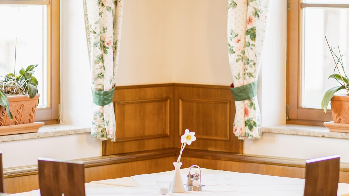 Ein gemütlicher Gastraum mit Holzstühlen und einem Tisch in der Ecke. Die Fenster sind mit floralen Vorhängen dekoriert, und an der Wand hängt ein Kreuz.