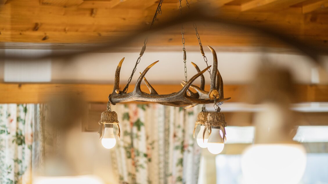 Ein rustikaler Kronleuchter aus Geweih hängt von der Holzdecke. Im Hintergrund sind Vorhänge mit floralem Muster sichtbar.