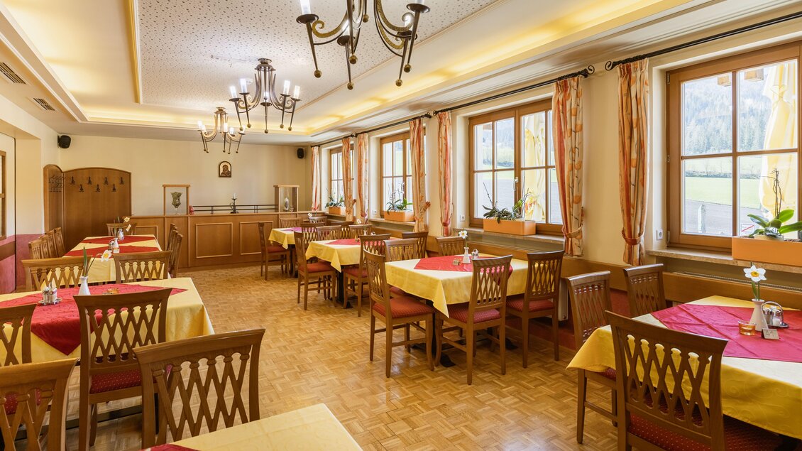 Ein einladendes Restaurant mit hellen Wänden und großen Fenstern. Die Tische sind mit roten und gelben Tischdecken gedeckt und bieten Platz für Gäste.