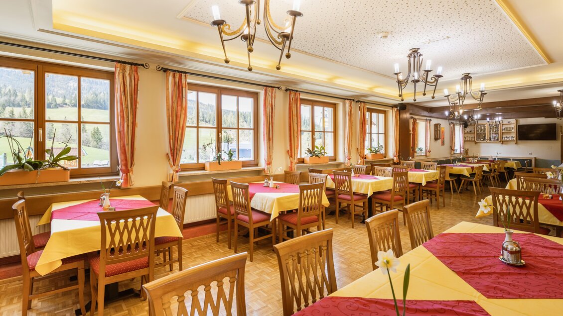 Ein gemütliches Restaurant mit Holzstühlen und Tischen in rot-gelber Tischdecke. Große Fenster bieten einen Blick auf die Natur.