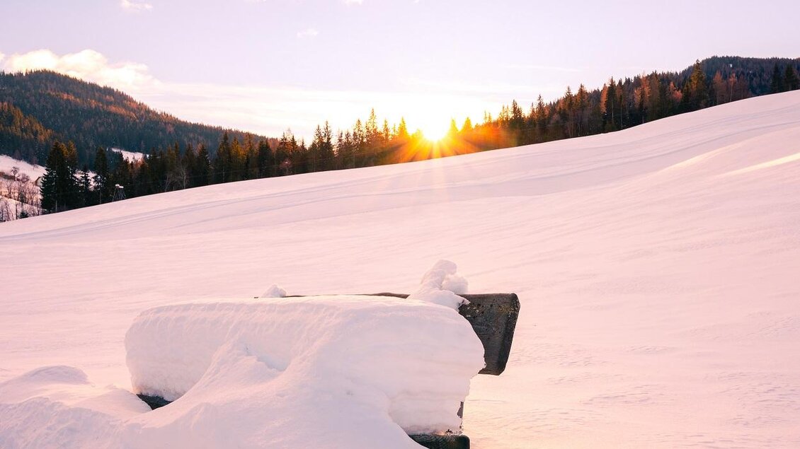 Eine verschneite Landschaft mit einer Bank, die teilweise im Schnee versteckt ist. Im Hintergrund ist die Sonne, die über den Hügeln aufgeht.