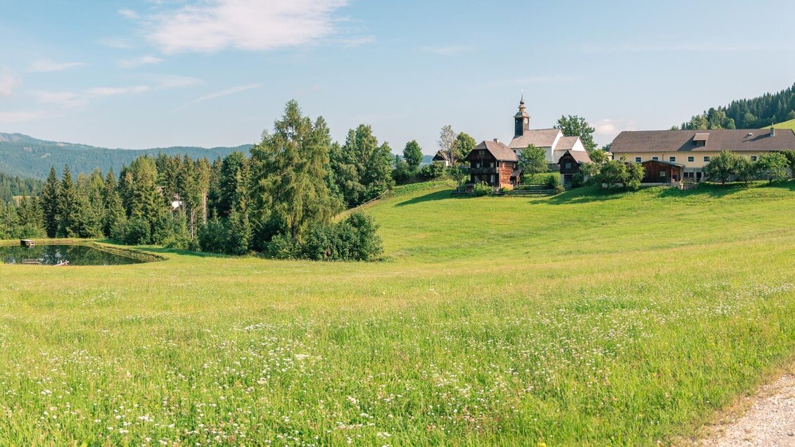 Eine idyllische Landschaft mit grünen Wiesen und sanften Hügeln. Im Hintergrund sieht man ein kleines Dorf und einen klaren Himmel.