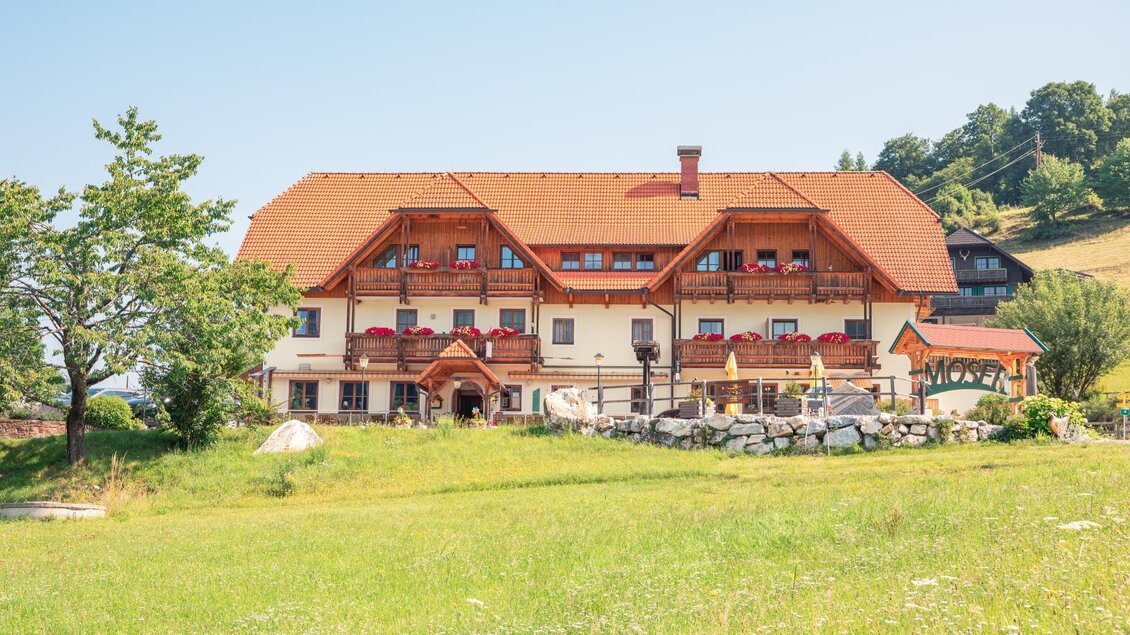 Ein traditionelles Haus mit rotem Dach in einer grünen Landschaft. Im Hintergrund sind sanfte Hügel zu sehen.