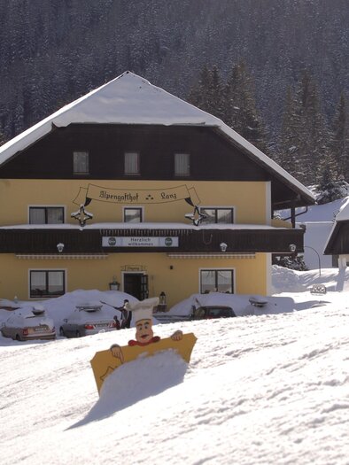 A yellow building in the snow, surrounded by a wintry landscape. The surroundings are covered in snow, and trees can be seen in the background. | © Alpenhotel Lanz