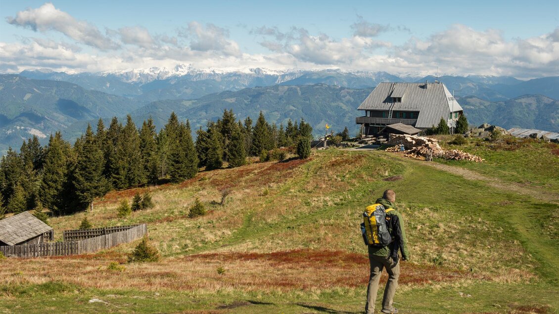 Ein Wanderer steht auf einer grünen Bergwiese und blickt auf eine Berghütte im Hintergrund. Die Landschaft ist von Wäldern und Bergen umgeben, unter einem klaren Himmel. | © TV Kapfenberg