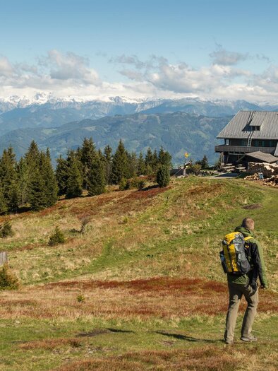 A hiker stands on a green mountain meadow, gazing at a mountain hut in the background. The landscape is surrounded by forests and mountains, under a clear sky. | © TV Kapfenberg
