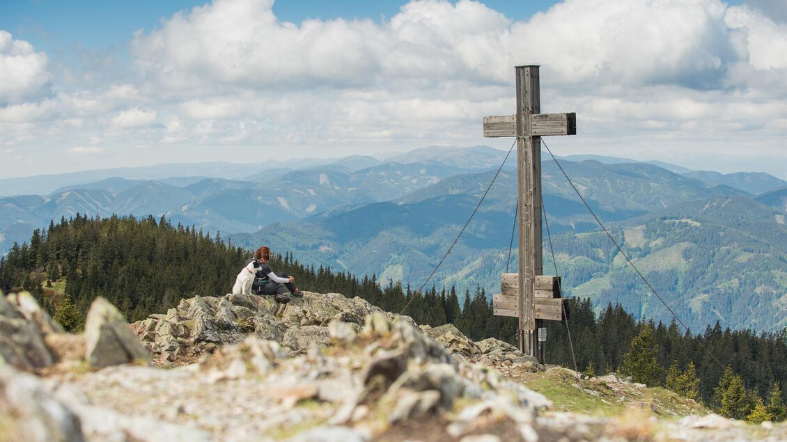 Ein Wanderer sitzt auf einem Felsen mit Blick auf die Berge. Im Hintergrund steht ein großes Holzkreuz unter einem bewölkten Himmel. | © TV Kapfenberg
