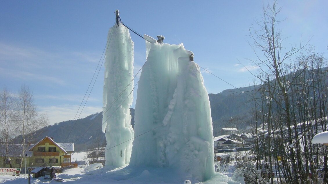 Ein beeindruckendes Eisgebilde neben einem Gebäude im Winter. Die Eisskulpturen ragen hoch in den blauen Himmel. | © Erlebnisregion Murtal