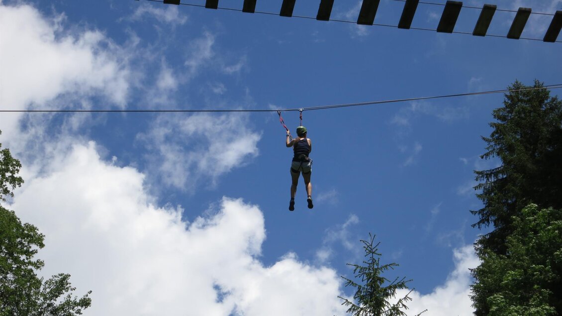 Ein Mensch schwebt an einem Seil über einem Wald. Im Hintergrund sind helle Wolken und Bäume zu sehen. | © Manuela Machner