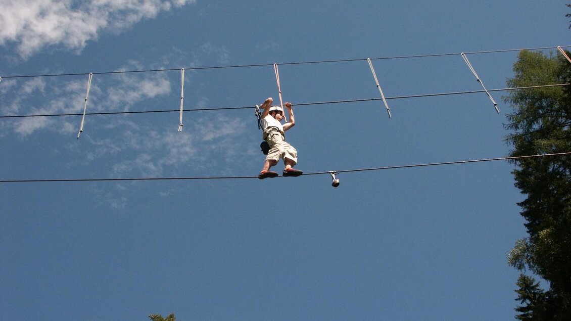 Ein Mensch balanciert auf einem Hochseil zwischen Bäumen. Der Himmel ist blau mit einigen Wolken. | © Ewald Walder