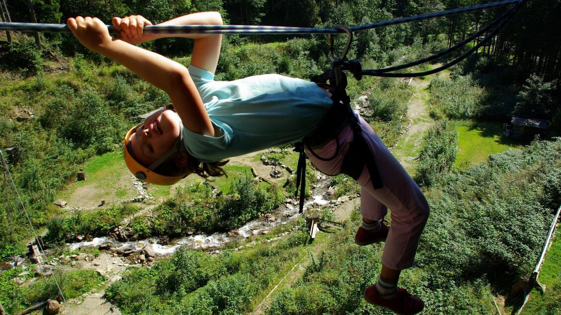 Ein Kind schwingt an einer Seilrutsche über einer grünen Landschaft. Im Hintergrund sind Bäume und ein fließender Bach zu sehen. | © Manuela Machner