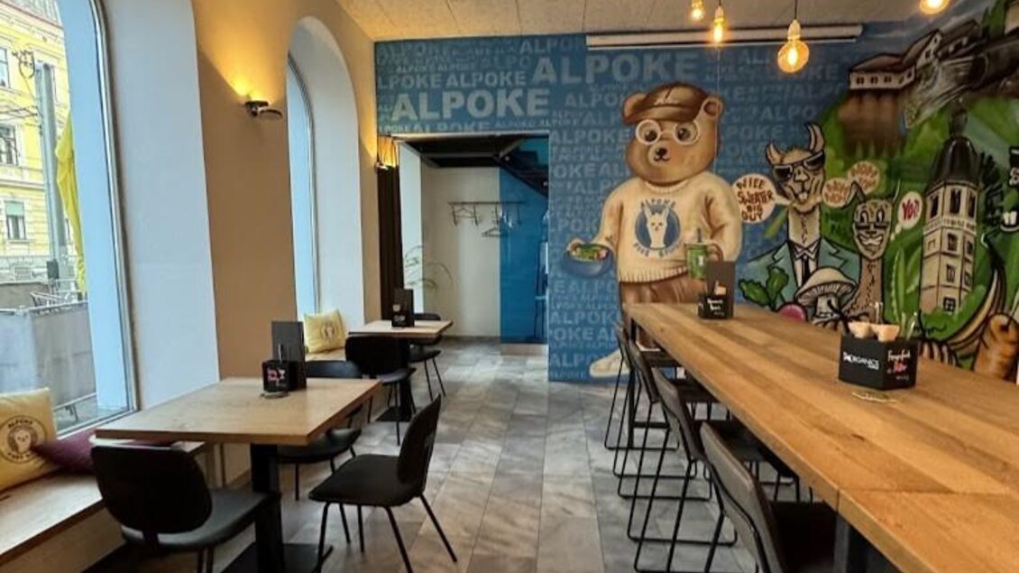 Ein modernes Café mit hellen Fenstern und Holzstühlen. An der Wand befindet sich eine bunte Wandmalerei eines Bären und anderen Figuren. | © Alpoke - K. van Kerken