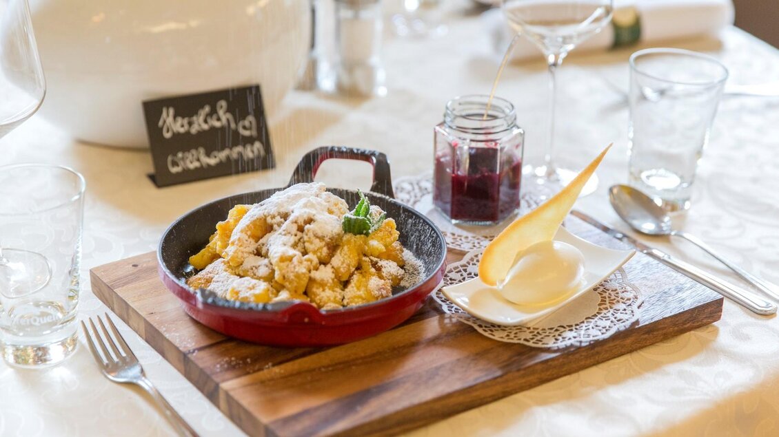 Ein köstliches Gericht in einer roten Pfanne, serviert mit einer weißen Creme und einer kleinen Schale Marmelade. Der Tisch ist elegant gedeckt mit Gläsern und Besteck. | © photoworkers