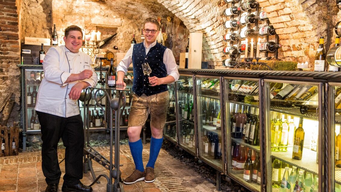Zwei Männer stehen in einem Weinkeller. Der Koch trägt eine weiße Uniform, während der andere in traditioneller bayerischer Kleidung mit einem Glas Wein posiert. | © photoworkers.at