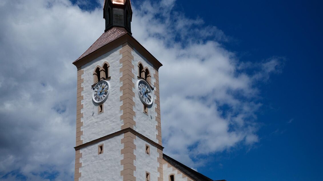 Eine Kirche mit einem hohen, spitzen Turm und einer kupfernen Dachspitze. Der Himmel ist blau mit einigen Wolken.