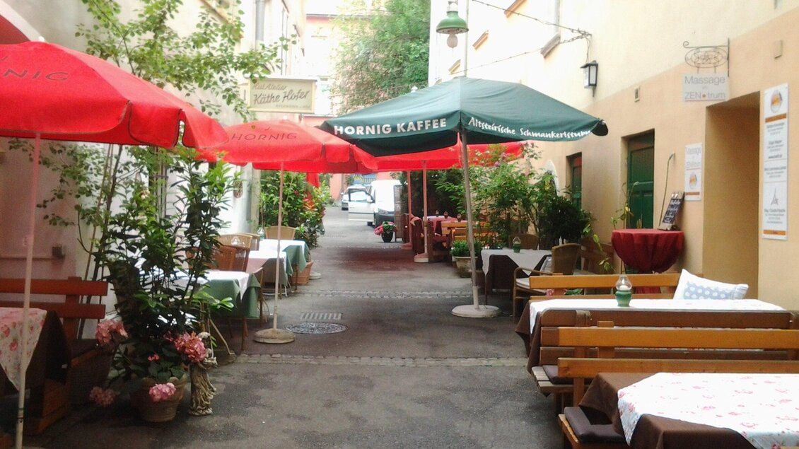 Eine ruhige Gasse mit Tischen und Stühlen unter bunten Schirmen. Pflanzen und Dekoration verleihen der Umgebung einen einladenden Charme. | © Altsteirische Schmankerlstube