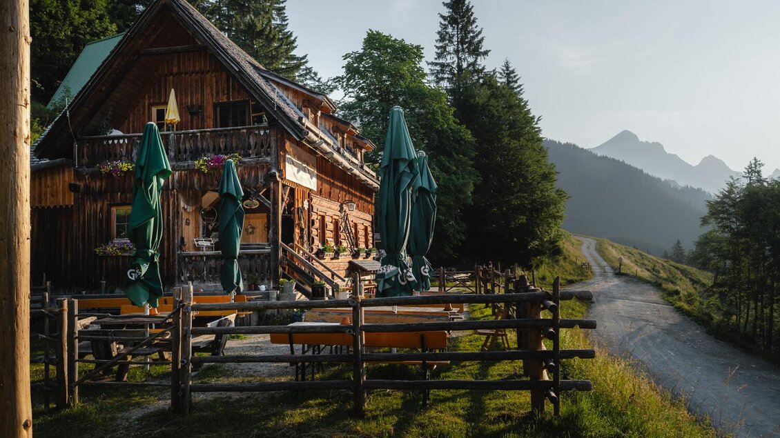 Ein traditionelles Holzhaus in den Bergen mit grünen Sonnenschirmen. Im Hintergrund sind hohe Bäume und bergige Landschaften zu sehen.