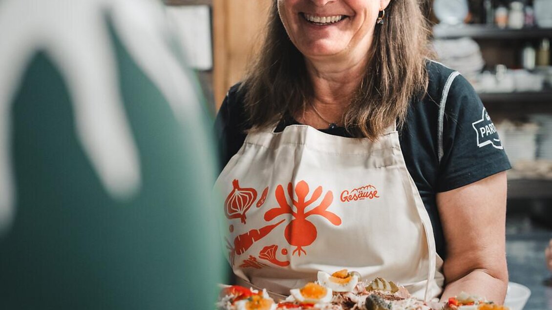 Eine lächelnde Frau trägt ein Tablett mit Essen. Sie steht in einer gemütlichen Küche mit Holzregalen im Hintergrund.