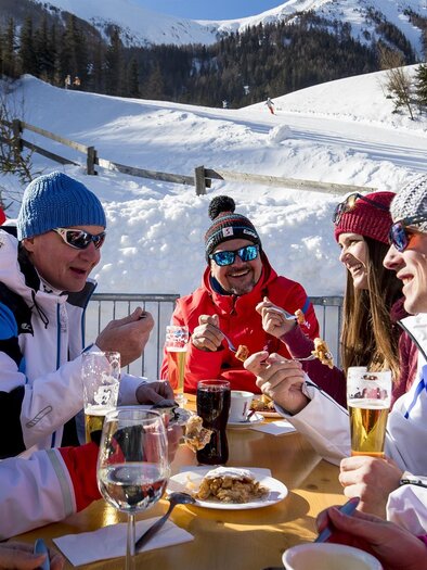 Eine Gruppe von Freunden genießt ein Essen im Freien in einer verschneiten Berglandschaft. Sie tragen warme Kleidung und lachen zusammen, während sie die winterliche Atmosphäre erleben. | © Präbichl Bergbahnen
