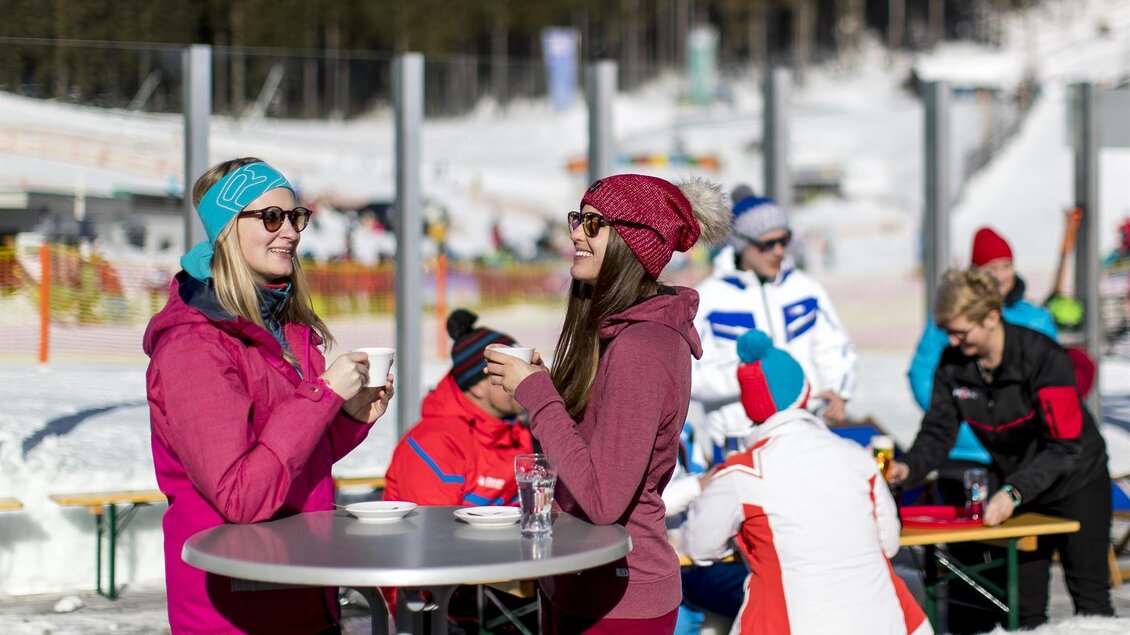 Zwei Frauen in warmer Winterkleidung unterhalten sich bei einem Tisch auf einer verschneiten Terrasse. Im Hintergrund sind weitere Personen und Skifahrer zu sehen. | © Präbichl Bergbahnen