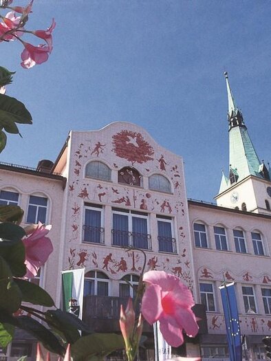 Ein historisches Gebäude mit einer grünen Turmspitze und bunten Fassadengemälden. Vorne blühende Pflanzen verleihen dem Bild eine frische Note. | © Robert Cescutti