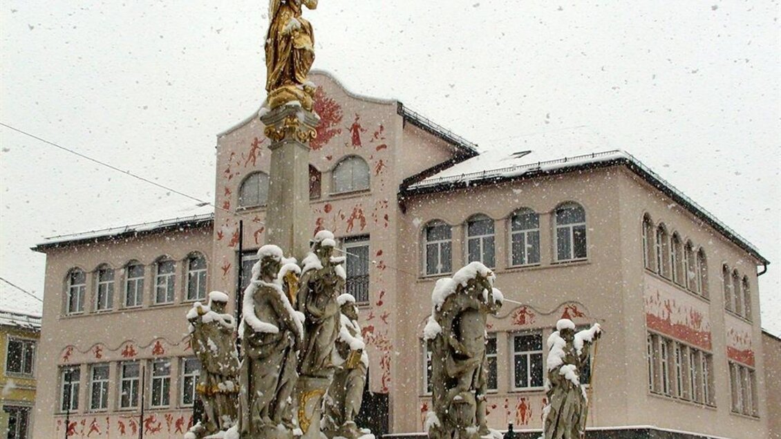 Ein historischer Platz mit einer Statue und mehreren Skulpturen, die mit Schnee bedeckt sind. Im Hintergrund ist ein Gebäude mit bunten Wandmalereien zu sehen. | © Robert Cescutti
