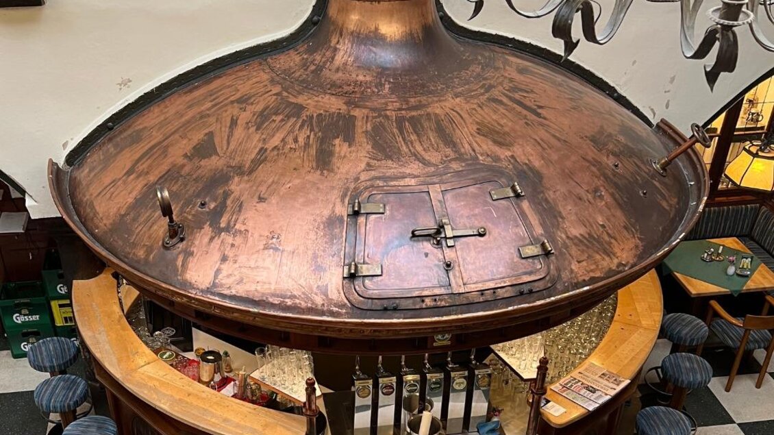 Eine elegante Bar mit einer großen, kupfernen Theke. Rund um die Bar stehen Hocker und die Umgebung ist gemütlich gestaltet. | © Arkadenhof