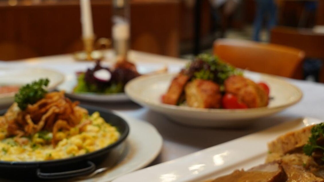 Ein schöner Tisch mit verschiedenen Gerichten, darunter Braten, Kartoffeln und Pasta. Im Hintergrund ist eine gemütliche Restaurantatmosphäre zu sehen. | © Arkadenhof