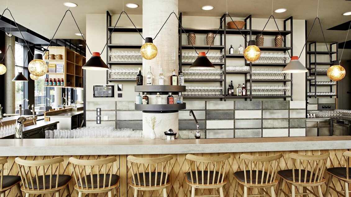 Eine moderne Bar mit Holzstühlen und einer eleganten Theke. Im Hintergrund sind Regale mit Gläsern und Getränken zu sehen. | © Arravané - Gerhard Wasserbauer