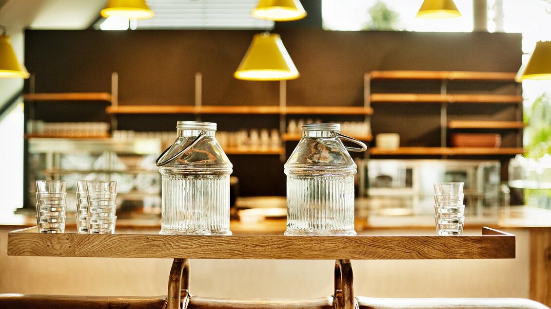 Ein stilvoll eingerichteter Raum mit einem Holztablett, das zwei Glaskrüge und mehrere Gläser hält. Der Hintergrund zeigt eine moderne Bar mit gelben Lampen. | © Arravané - Gerhard Wasserbauer