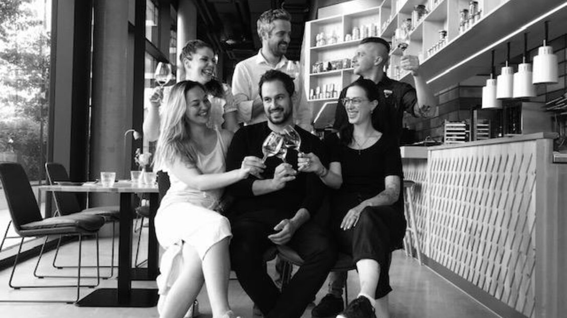 Eine Gruppe von sechs Menschen steht und sitzt in einem modernen Café. Sie stoßen mit Gläsern an und lachen gemeinsam. | © Arravané