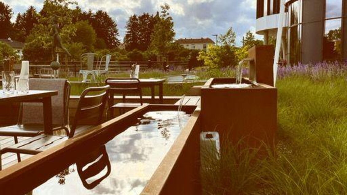 Ein moderner Außenbereich mit Gartenmöbeln und einer Wasserfläche reflektiert die Wolken. Im Hintergrund sind Bäume und ein modernes Gebäude sichtbar. | © Arravané