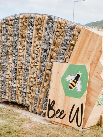 An innovative bee house made of wood and stones, standing on a green lawn. It has a modern design with a bee symbol and an inviting appearance. | © MotionAds