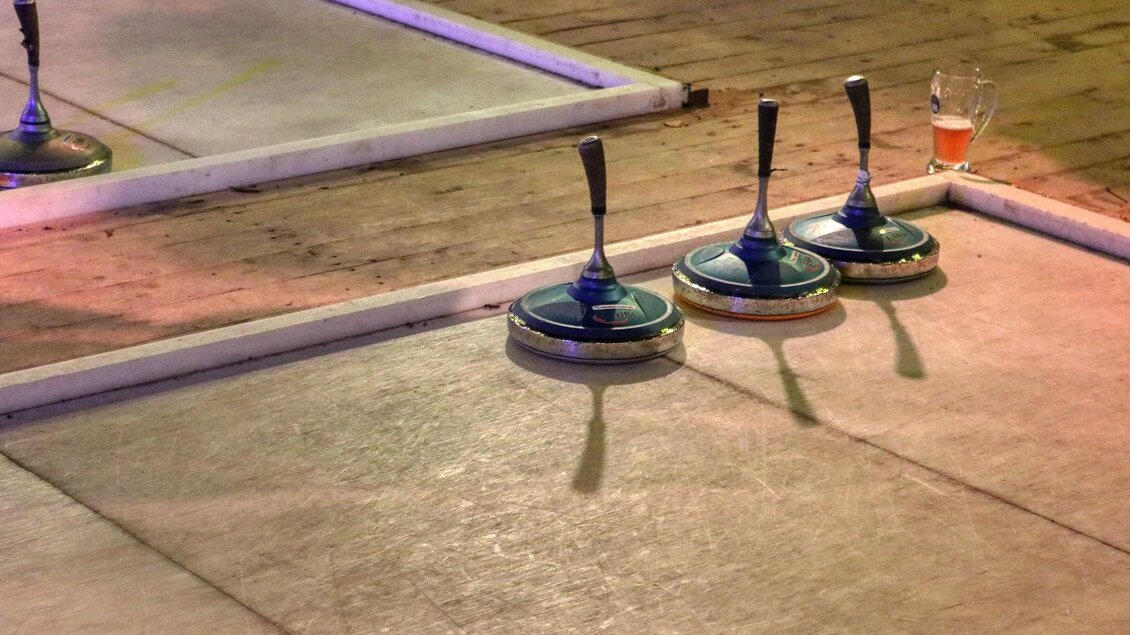 Eine Curlingbahn mit drei Curlingsteinen und einem Getränk im Hintergrund. Die Umgebung ist beleuchtet und schafft eine entspannte Atmosphäre. | © Pixabay