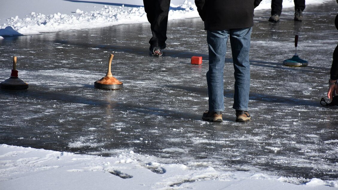 Eine Gruppe von Menschen spielt auf einer gefrorenen Fläche mit Curling-Steinen. Die Umgebung ist mit Schnee bedeckt, und es herrscht sonniges Wetter. | © Eisstockschießen Gasthaus Jagawirt