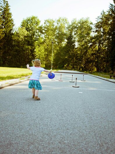 A little girl happily runs on a street with green trees in the background. In front of her are some set-up objects for playing. | © Häuserl im Wald 