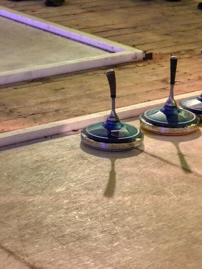 Three curling stones on a lane, surrounded by a wooden area. A drink is visible in the background. | © Pixabay