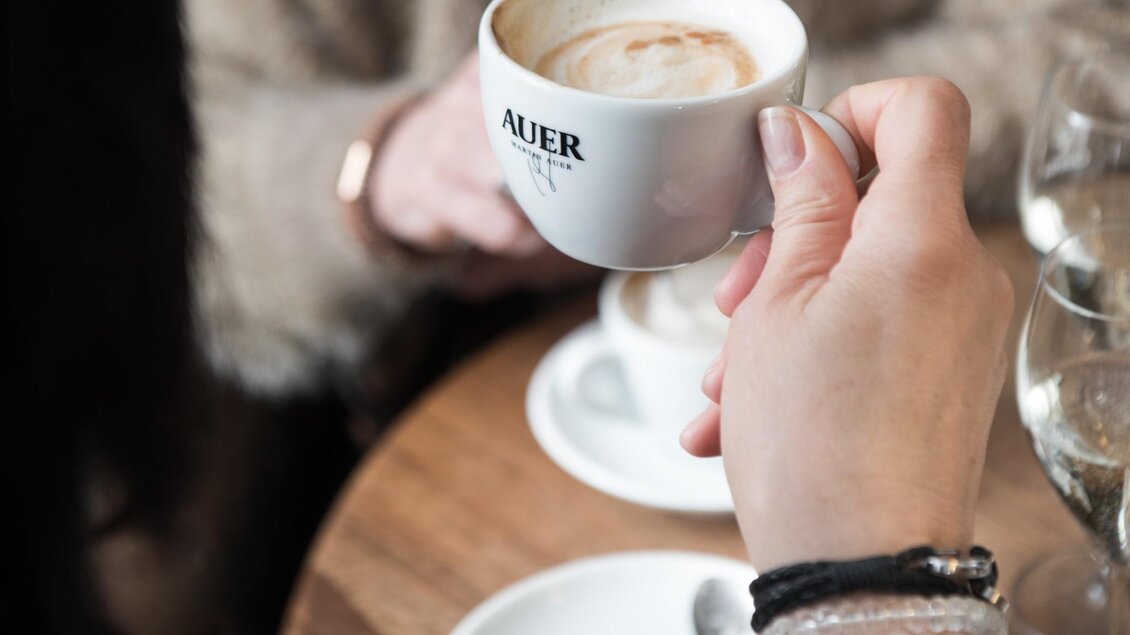 Hand hält Cappuccino-Tasse mit Martin-Auer-Logo beim Kaffee mit Freundinnen. | © Martin Auer