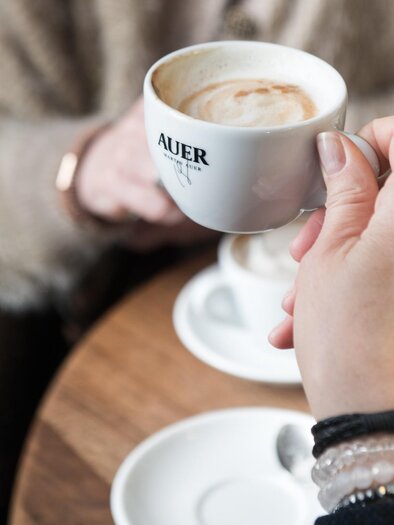 Hand hält Cappuccino-Tasse mit Martin-Auer-Logo beim Kaffee mit Freundinnen. | © Martin Auer